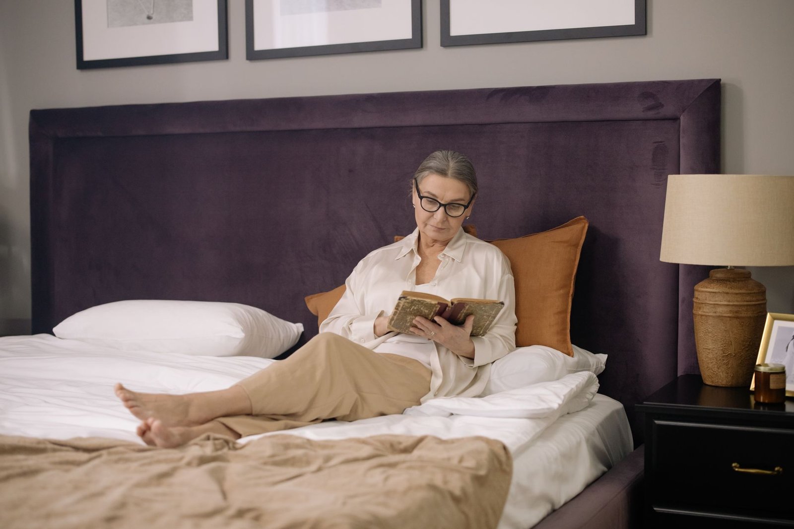 slaapproblemen 50 plus — A senior woman enjoys reading a book in a cozy and elegant bedroom setting.